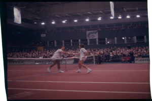 Zwei Männer spielen Tennis auf einem Platz mit einem Netz, die Rackets haltend, vor einer großen Menge mit Menschen auf den Tribünen und Bannern im Hintergrund.