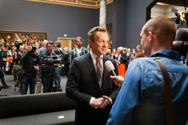 Ein Mann im Anzug und Krawatte spricht mit einem Reporter vor einer Menge, einige halten Kameras, mit einem Sofa im Vordergrund und einer Wand mit einem Foto Rahmen, Lampen und einer Säule im Hintergrund, während er ein Mikrofon hält.