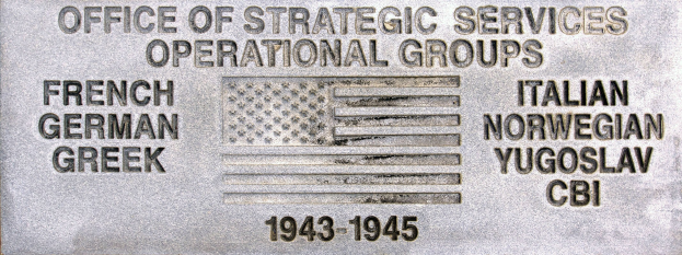 Metallschild an einer Gebäudeseite mit der Aufschrift "Office of Strategic Services Operational Groups" und einer Flagge im Hintergrund, mit eingravierten Text und Zahlen.