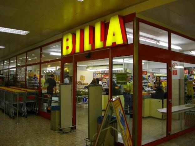Ein Supermarkt mit einer mittigen Eingangstür, Mülltonnen daneben, einer Reihe Einkaufswagen auf der linken Seite und einigen Menschen drinnen, die Produkte anschauen und herumlaufen, mit Licht an der Decke.