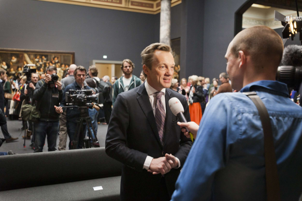 Ein Mann im Anzug hält ein Mikrofon und wird von einem Reporter vor einer Menge mit Kameras interviewt, ein Sofa im Vordergrund, ein Rahmen an der Wand und Beleuchtungsausrüstung.