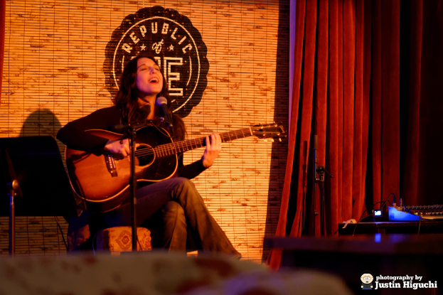 Eine Person sitzt auf einem Stuhl und spielt eine Gitarre und singt in ein Mikrofon, mit einer Vorhangwand dahinter, verschiedenen Instrumenten in der NÃ¤he und einem Notenhalter vorne.