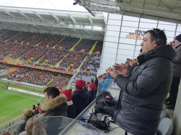 Ein Mann in einer Jacke und Kopfhörern steht vor einer Menge bei einem Fußballspiel, mit Taschen und Gegenständen auf einem Tisch im Vordergrund und einem Stadion mit Zuschauern im Hintergrund.