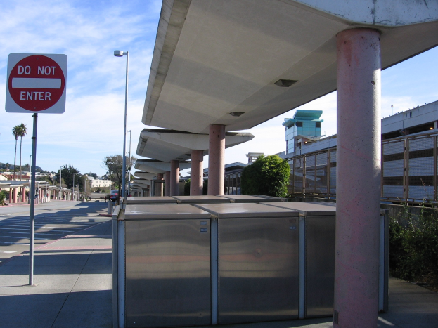Eine Bushaltestelle mit einem "Betreten Verboten"-Schild an der Straße, mit Metallcontainern, Pfählen, Lampen, Bäumen, Pflanzen und Gebäuden im Vordergrund und einem bewölkten Himmel im Hintergrund.