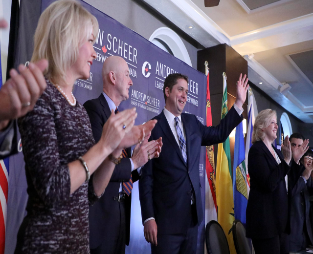 Eine Gruppe von Menschen, die vor Fahnen stehen und klatschen, mit einem Tisch voller Gegenstände rechts daneben, Stühlen, einem Banner und einer Wand im Hintergrund sowie Lichtern und einem Ventilator auf dem Dach.