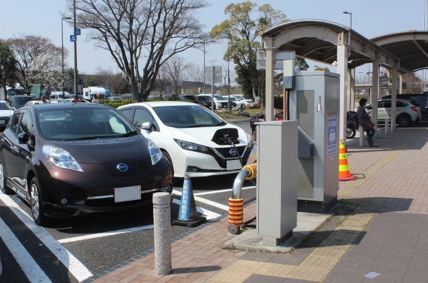 Elektrisches Auto-Ladestation in Japan mit Autos auf der Straße, Verkehrskegel, einer Person auf dem Gehweg, einem Schuppen, Masten, Lichtern, Schildern, Bäumen, Pflanzen und einem Himmel im Hintergrund.