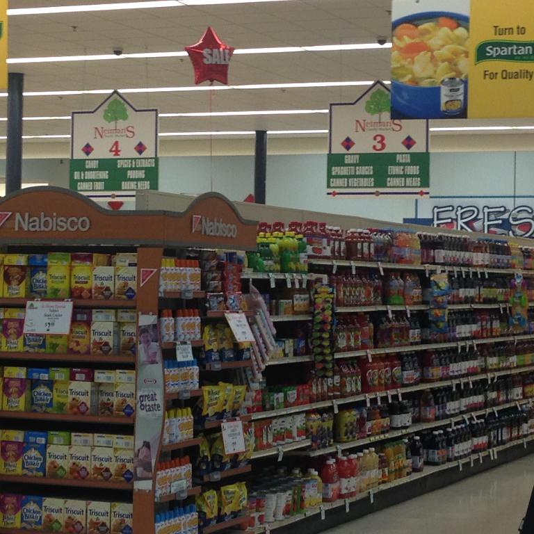 Ein Supermarkt-Innenraum mit Regalen voller Lebensmittel, Deckenlampen und Schildern.