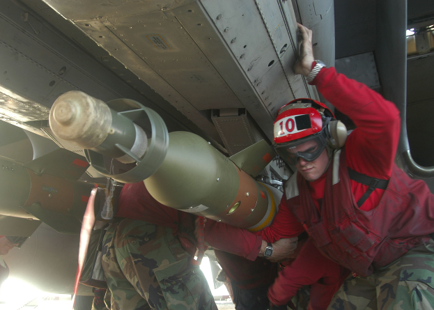 Eine Gruppe von M"ännern in roten Jacken und Tarnuniformen arbeitet an einem Flugzeug, wobei einer einen Helm, eine Schutzbrille und eine Uhr tr"agt und im Hintergrund eine Rakete zu sehen ist.