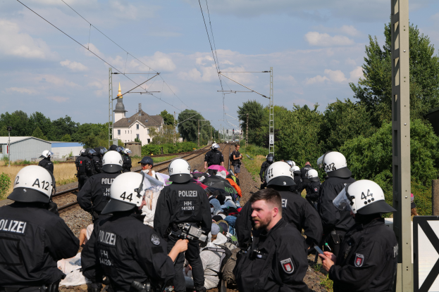 Eine Gruppe von Polizisten in schwarzen Uniformen und weißen Helmen, einige bewaffnet, steht auf Gleisen mit Bäumen, Pfählen, Drähten, Gebäuden und einem klaren blauen Himmel im Hintergrund.