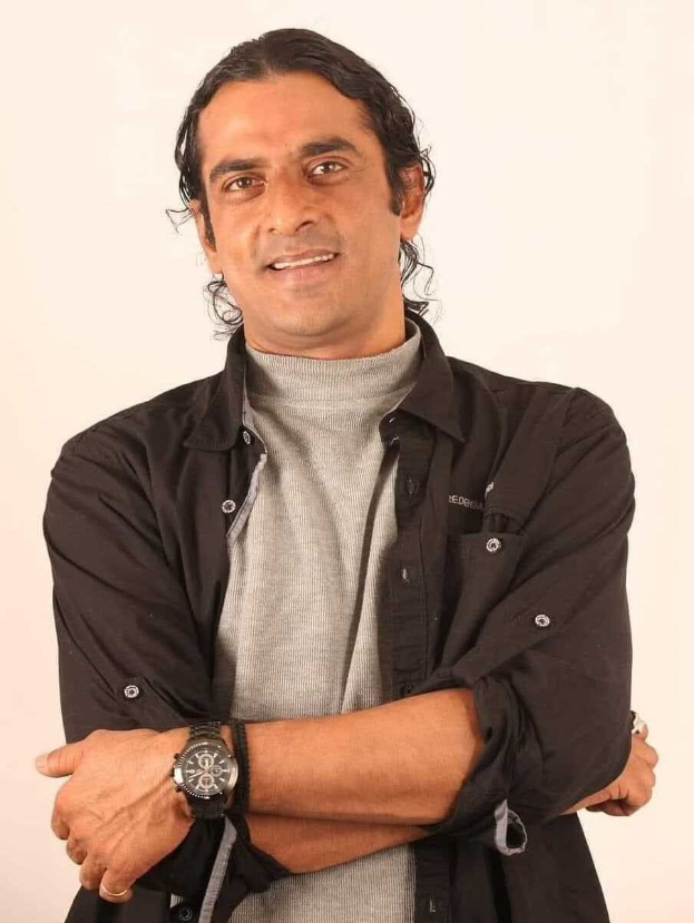 Ein Mann in einem schwarzen und grauen Outfit mit überschlagenen Armen und einer Uhr, der vor einem wei├čen Hintergrund steht und nachzudenken scheint.