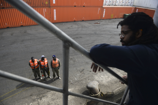 Ein Mann in legerer Kleidung steht auf einem Metallgelönder in der Nähe einer Gruppe von Menschen in orangefarbenen Jacken und Kappen, mit Containern und einer Wand im Hintergrund am Hafen von Los Angeles.