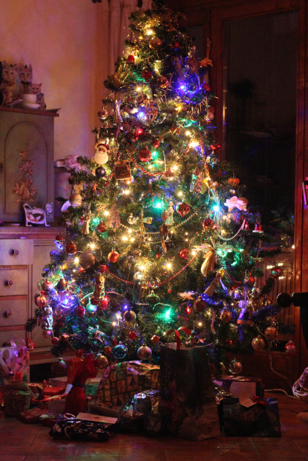 Ein Weihnachtsbaum mit wenigen Lichtern und Dekorationen, Geschenke auf dem Boden, ein Schrank mit Gegenständen links und ein Fenster an der rechten Wand.