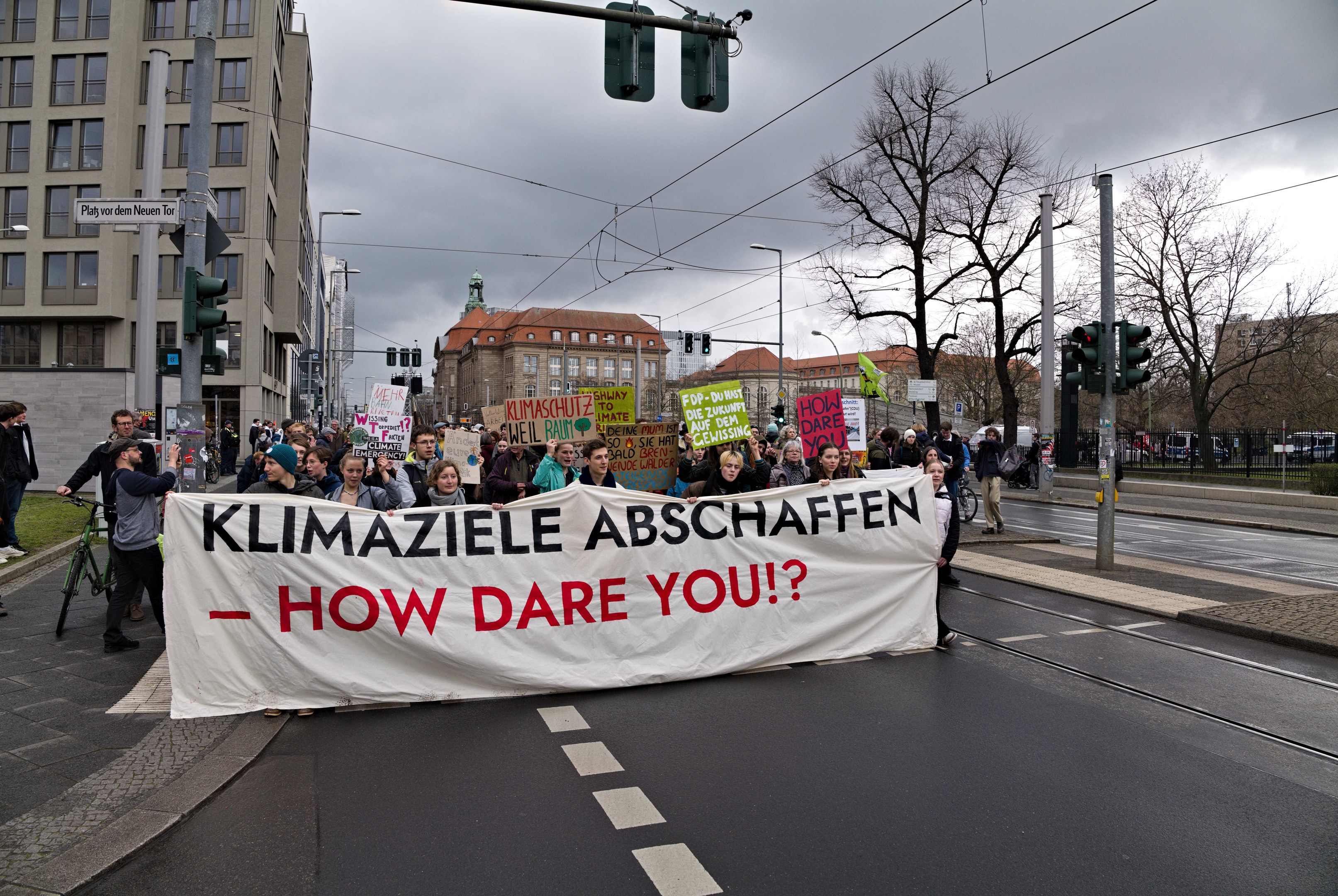 Eine Gruppe von Menschen, die eine Straße entlanggehen und ein Banner mit der Aufschrift "Klimaziele Abschaffen - Wie wagst du es?" tragen, mit einem Radfahrer im Vordergrund und Gebäuden, Bäumen, Verkehrssignalen und einer klaren blauen Himmel im Hintergrund.