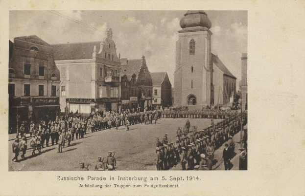 Ein Schwarz-Weiß-Foto eines Umzugs in Insterburg am 5. September 1914 mit vielen Menschen, Gebäuden im Hintergrund und Text am unteren Bildrand.