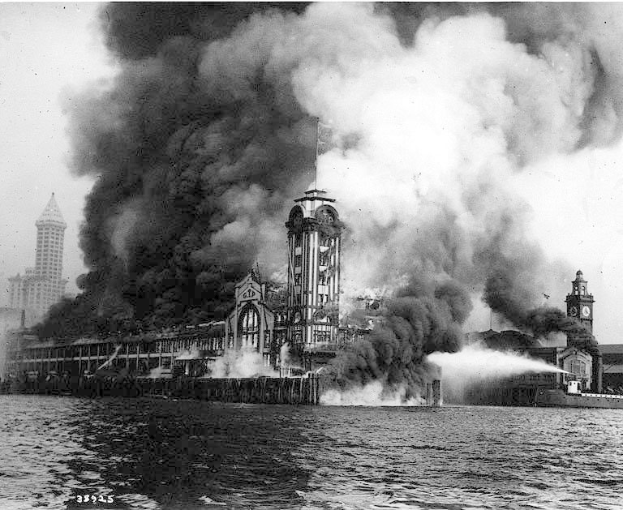 Schwarzes und weißes Foto eines großen Gebäudes, das in Flammen steht und aus dem Rauch aufsteigt, mit einem Gewässer im Vordergrund und anderen Gebäuden und dem Himmel im Hintergrund.