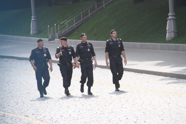 Drei Polizisten in Uniform und Mützen, die eine Straße mit Geländern, Treppen und Gras im Hintergrund entlanggehen.