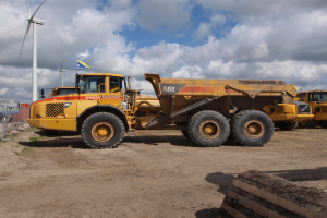 Ein großer gelber Muldenkipper steht vor einer Windturbine, umgeben von anderen Fahrzeugen und einem Himmel voller weißer, flauschiger Wolken im Hintergrund.