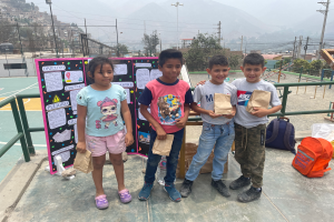 Eine Gruppe von Kindern steht auf einem Basketballfeld, jedes hält einen Papiertüte, mit Tüten und Gegenständen rechts daneben und einer Tafel mit Text links daneben, mit Zaun, Pfosten, Straßenlaternen, Strommasten mit Drähten, Bäumen, Gebäuden, Hügeln und Himmel im Hintergrund.