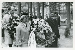Eine Gruppe von Menschen steht nebeneinander, jeder hält einen Blumenstrauß in der Hand, auf einem Schwarz-Weiß-Bild mit Bäumen und Gebäuden im Hintergrund, wahrscheinlich bei einer Holocaust-Gedenkfeier.