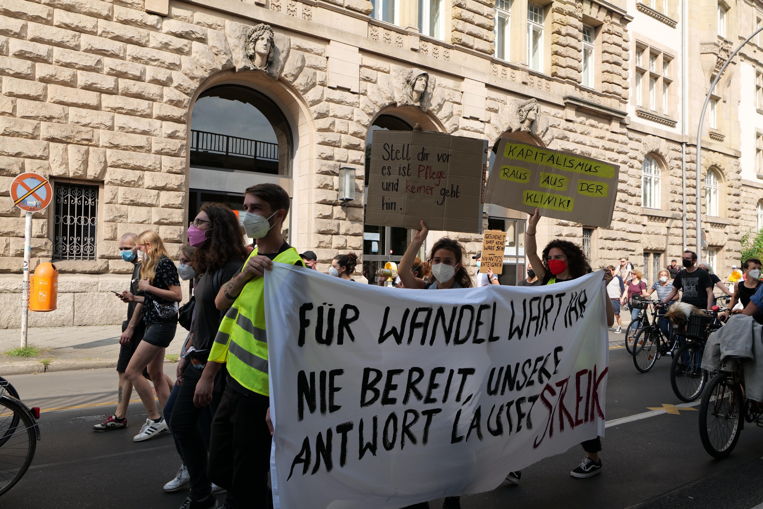Eine Gruppe von Menschen marschiert bei einer Protestaktion auf einer Stadtstraße, einige halten Schilder und andere fahren Fahrräder, im Hintergrund ein historisches Gebäude mit Rundbogenfenstern, Säulen und Skulpturen.