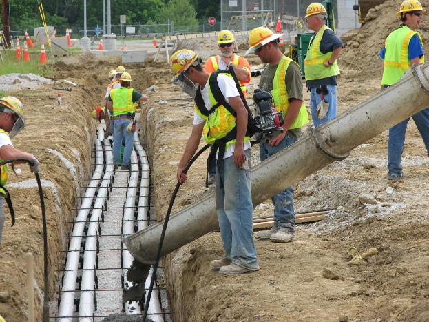 Eine Baustelle mit Arbeitern in Jacken und Helmen, Absperrgitter, Zaun, Bäume, Pfosten, Erde und Rohre im Hintergrund.