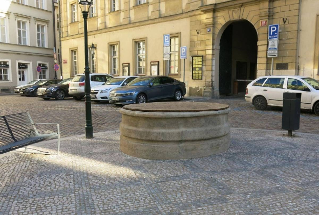 Ein Stadtplatz mit einer Bank, einem Brunnen, Laternenmasten und Schildern, umgeben von Gebäuden mit Fenstern.