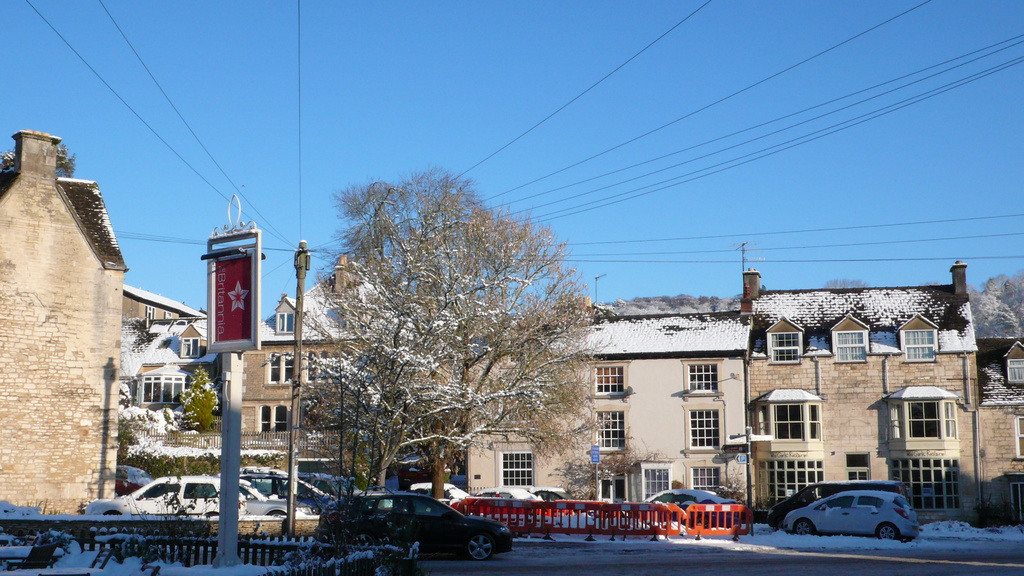 Fahrzeuge auf einer schneebedeckten Straße mit Häusern, Bäumen und Strommasten im Hintergrund, alles von Schnee bedeckt.