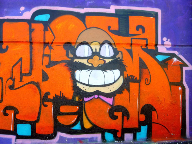 Graffiti-Gemälde eines Cartoon-Charakters mit großen Augen, einem breiten Lächeln und einem verschmitzten Gesichtsausdruck an der Seite eines Gebäudes, mit leuchtenden Farben und auffälligem Begleittext.