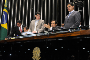 Leute sitzen an einem Tisch auf einer Bühne mit einer Holzpaneelwand und Fahnen dahinter und einem goldenen Logo auf dem Tisch vorne.