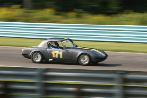 Ein Auto mit einem Helm tragenden Fahrer fährt auf einer Straße entlang, die von Barrieren gesäumt ist und Bäume im Hintergrund hat.