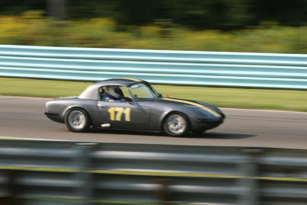Ein Auto mit einem Helm tragenden Fahrer fährt auf einer Straße entlang, die von Barrieren gesäumt ist und Bäume im Hintergrund hat.