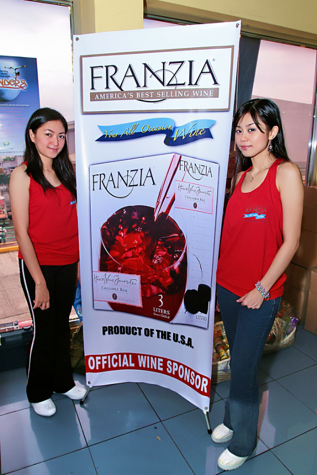 Zwei Frauen neben einem Plakat für Franzia, dem bestverkauften Wein Amerikas, mit einem Glasfenster im Hintergrund.