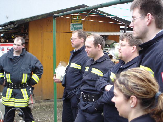 Eine Gruppe von Männern in blauen Feuerwehrausrüstungen, die nach links schauen, mit einem kleinen Holzschuppen und Rohrrahmen im Hintergrund.