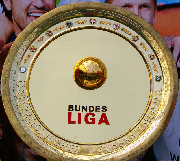 Ein Mann mit einem goldenen Schild mit der Aufschrift 'Bundesliga', im Hintergrund eine Fahne mit drei Personen.