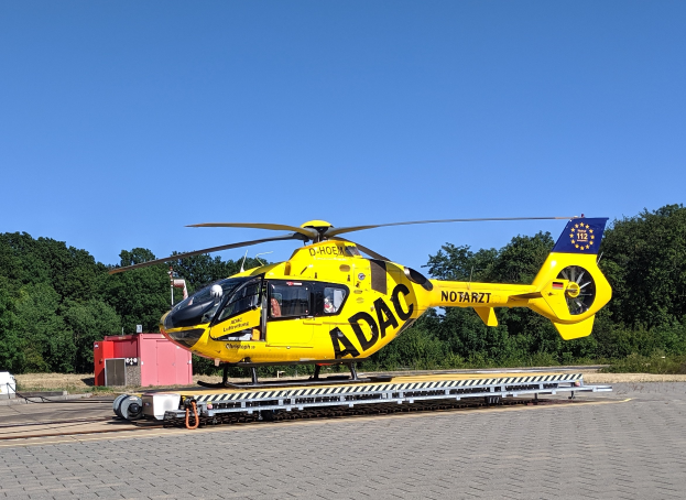 Ein gelber Hubschrauber steht auf einem Flughafen-Asphalt, umgeben von Bäumen und einem klaren blauen Himmel, mit verschiedenen Gegenständen, die mit dem Hubschrauber in Zusammenhang stehen, herum verstreut.