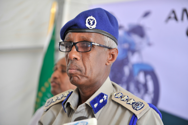 Ein Mann in Polizeiuniform steht an einem Podium mit Mikrofonen, trägt eine Mütze und Brille und hat eine weitere Person, eine Fahne und ein Banner im Hintergrund.