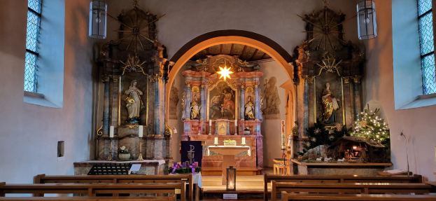 Das Innere einer Kirche mit hölzernen Kirchenbänken, Kerzen, Blumenbouquets, Statuen und einem geschmückten Weihnachtsbaum im Hintergrund.