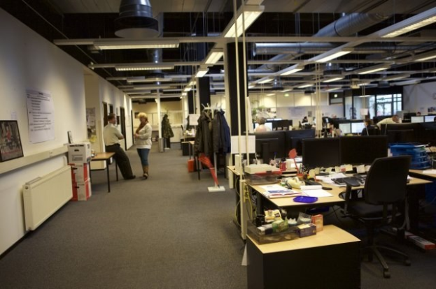 Großes Großraumbüro mit Menschen, die an Schreibtischen mit Computern, Monitoren, Tastaturen und anderen Büromaterialien arbeiten, mit aufgehängten Kleidungsstücken, Wandtafeln mit Text und Bildern, Deckenleuchten und Glasfenstern im Hintergrund.