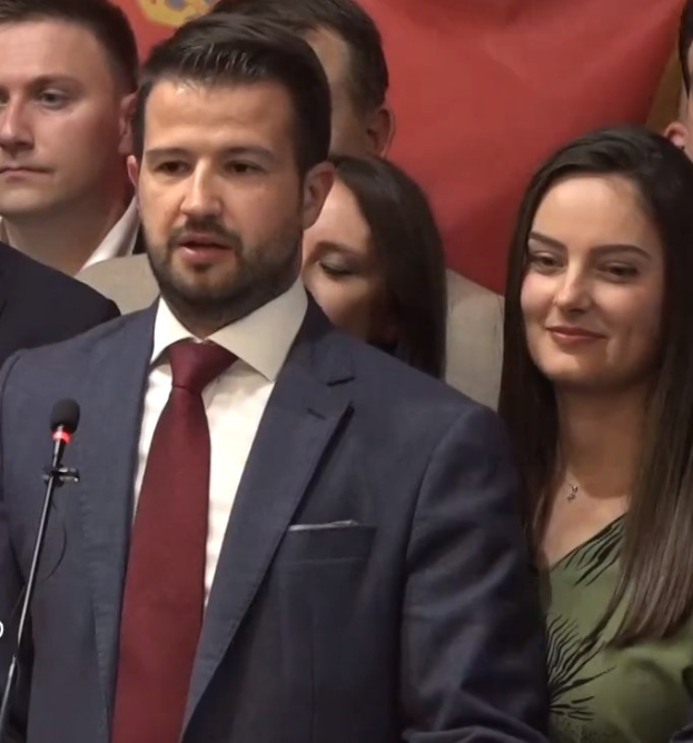 Ein Mann im Anzug spricht an einem Rednerpult mit einem Mikrofon zu einer Menge hinter einem Banner, wahrscheinlich über ein Regierungsozialmedienverbot.