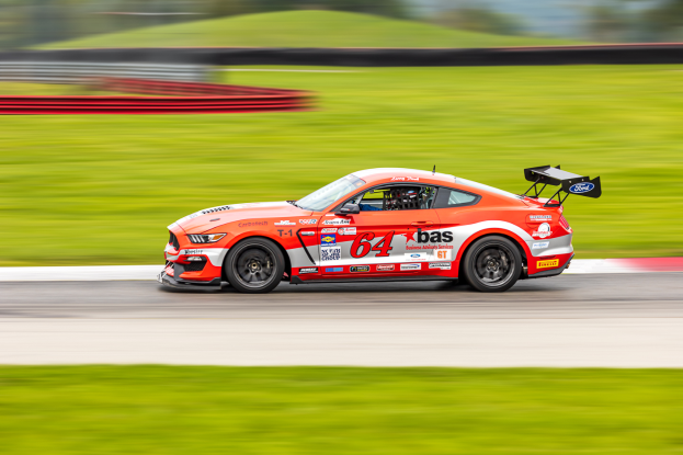 Ein Ford Mustang GT Rennauto, verziert mit Text und Zahlen, fährt auf einer Rennstrecke mit einem Helmträger am Steuer, Hintergrund unscharf, um Geschwindigkeit und Bewegung anzudeuten.