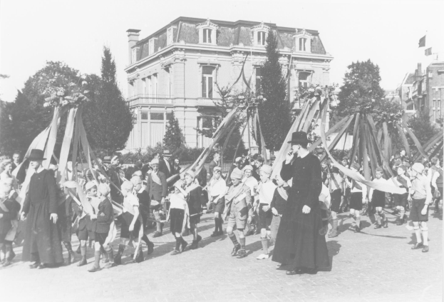 Ein Schwarz-Weiß- Bild einer Gruppe von Menschen, die eine Straße entlanggehen, einige halten Fahnen, mit Hüten, die einen Maibaum vor Gebäuden und Bäumen unter einem sichtbaren Himmel passieren.