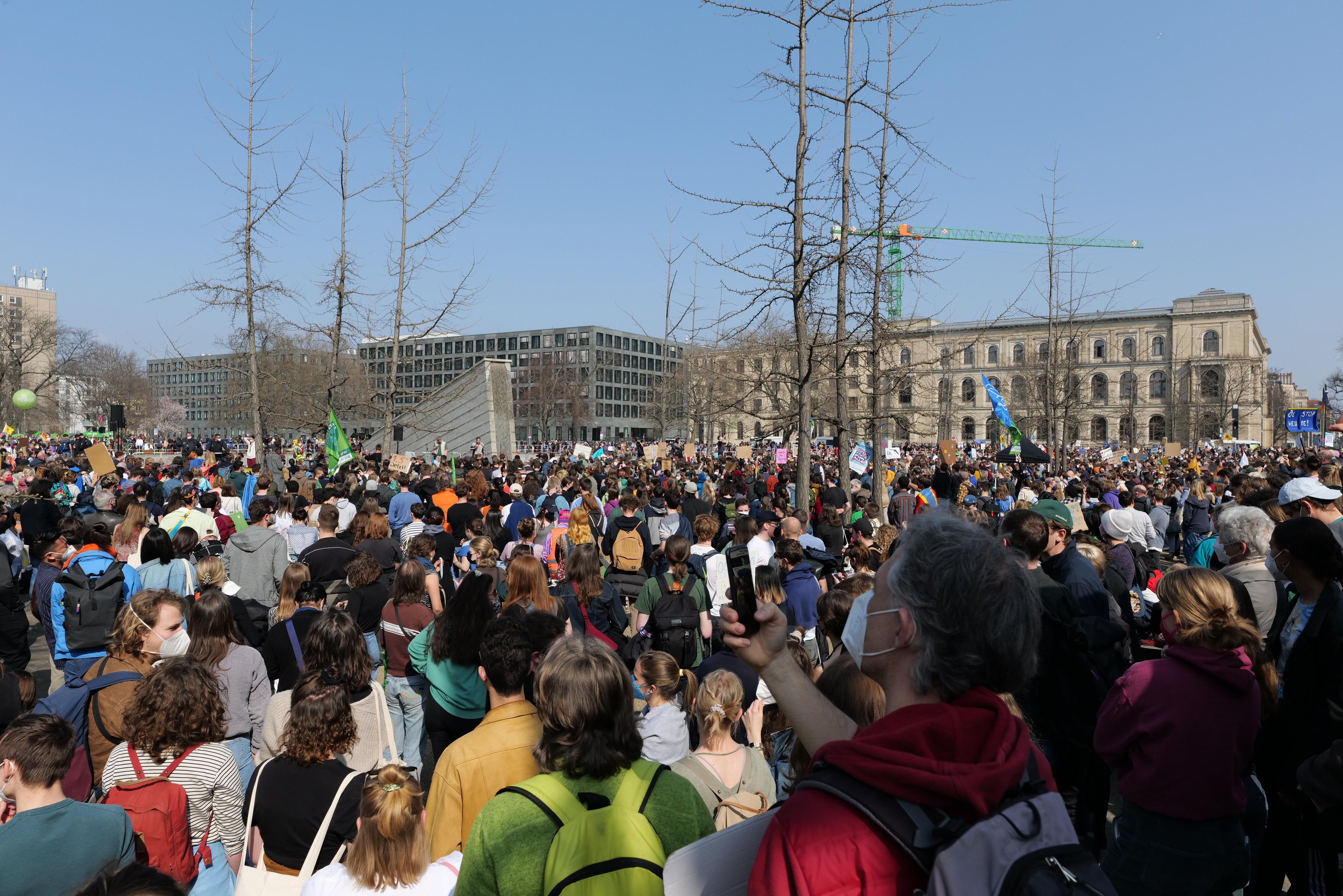 Große Menschenmenge mit Schildern und Taschen vor einem Gebäude mit Bäumen und einem klaren Himmel, was auf eine Klimawandel-Demonstration hinweist.