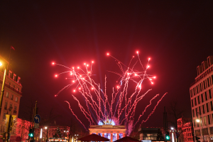 Eine Straßenansicht in Berlin an Silvester, voller Gebäude, Bäume, Laternenpfähle, Verkehrszeichen, Schilder, Zelte und Menschen, mit einem von Feuerwerk erleuchteten Himmel im Hintergrund.