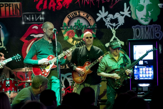 Drei Männer auf einer Bühne, die Gitarren spielen, mit einer Menge unten und einer bemalten Wand im Hintergrund.