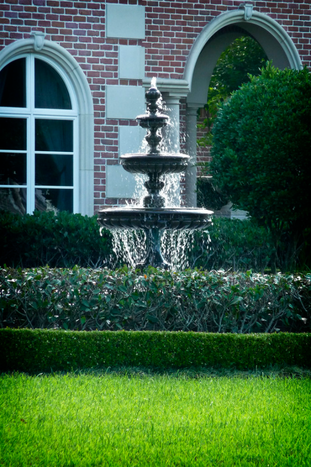 Eine GartenSzene mit Gras am Boden, einem zentralen Brunnen umgeben von ein paar Pflanzen und einem Gebäude im Hintergrund.