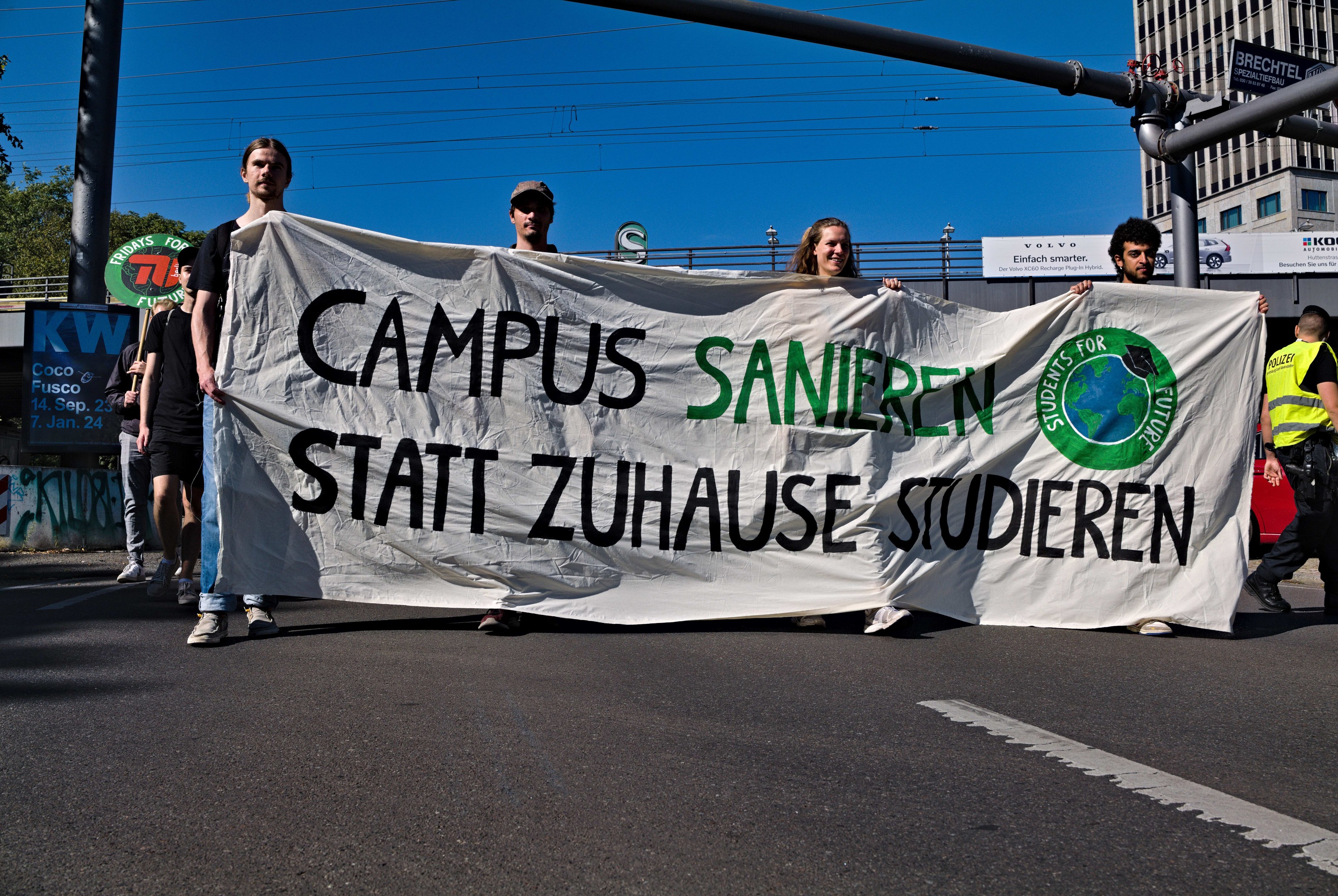 Gruppe von Menschen, die eine Straße entlanggehen und ein Transparent mit der Aufschrift "Campus Sanien Statt Zuhause Studieren" tragen, mit Gebäuden, Bäumen, Polen, Drähten und einem klaren blauen Himmel im Hintergrund.