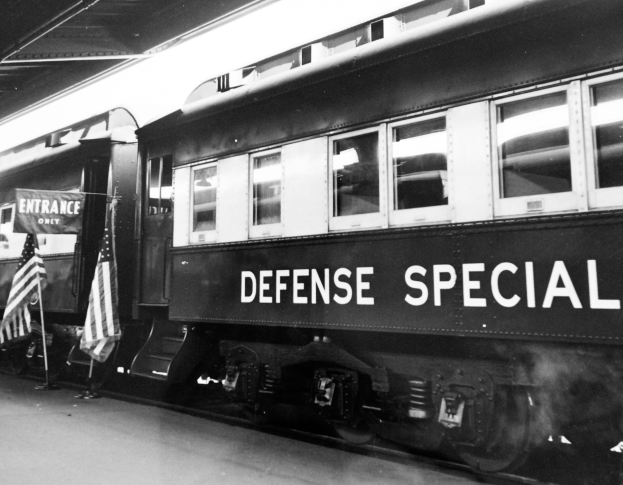 Ein Schwarz-Weiß-Foto eines Zuges mit der Aufschrift "Defense Special", der auf den Eisenbahnschienen geparkt ist, mit zwei Fahnen auf der linken Seite und einer Decke darüber.