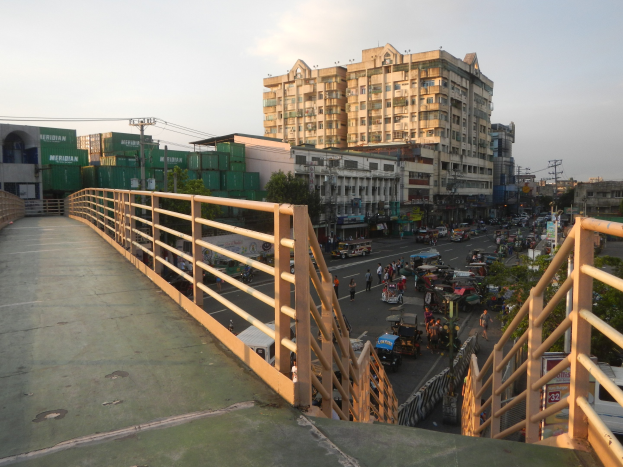 Fußgängerbrücke mit Geländern überspannt eine vielbefahrene Straße mit Fahrzeugen und Fußgängern, mit Gebäuden, Bäumen, Strommasten mit Drähten und einem bewölkten Himmel im Hintergrund.