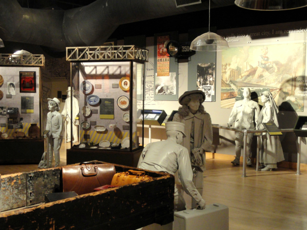 Ein Museumsraum mit verschiedenen Ausstellungsstücken, darunter Puppen mit Hüten, einer Aktentasche, einer Kiste und anderen Gegenständen auf dem Boden sowie Informationsschildern an den Wänden und Lampen an der Decke.