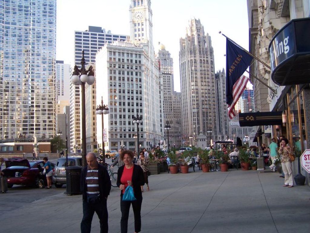 Eine belebte StraßenSzene mit Menschen, Blumentöpfen, Autos, Straßenlaternen, Gebäuden, Geschäften und Fahnen im Hintergrund.